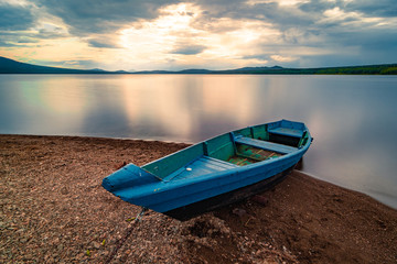 Blue wooden boat moored on the shore of lake under cloudy sky at sunset