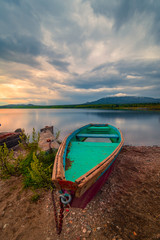Green wooden boat moored on the shore of lake under cloudy sky at sunset