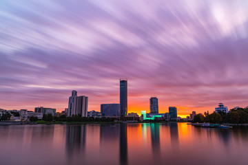 Beautiful purple and orange cloudy sunset at the city pond. Long Exposure cityscape of Yekaterinburg, Russia with skyscrappers reflecting in water