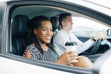 African young woman smiling at camera while drinking coffee during her trip by car together with young man