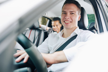 Portrait of young man smiling at camera while driving a car with female passenger in back seat
