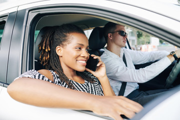 Happy African woman smiling and talking on mobile phone while travelling by car with man sitting near her and holding the wheel