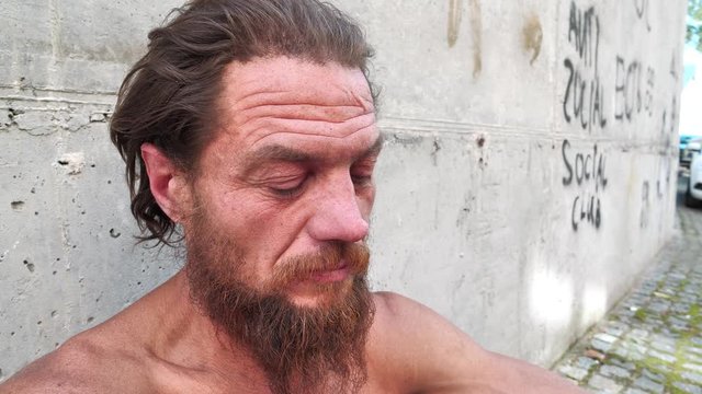 Destitute homeless man sits on the street and looks at camera. Real down-and-out person with no fixed abode