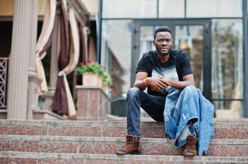 Young attractive serious african american man model. Modern handsome guy in jeans jacket sitting on urban city street.