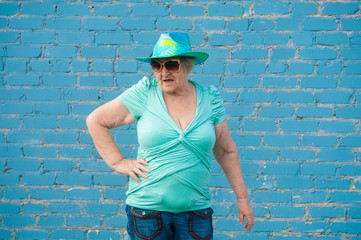 elderly grandmother in turquoise sweater and cowboy hat stands against gray brick wall