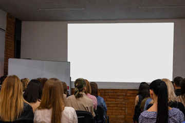 People in auditory during presentation or seminar. Teenagers or young men and women at university lecture or seminar. Back side view with no faces. There a big screen mock-up in front of them