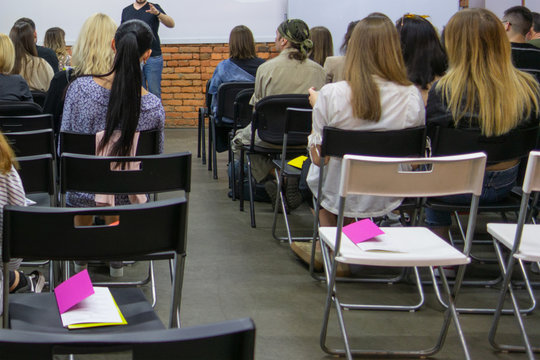 People in auditory during presentation or seminar. Teenagers or young men and women at university lecture or seminar. Back side view with no faces. There are empty chairs with booklets