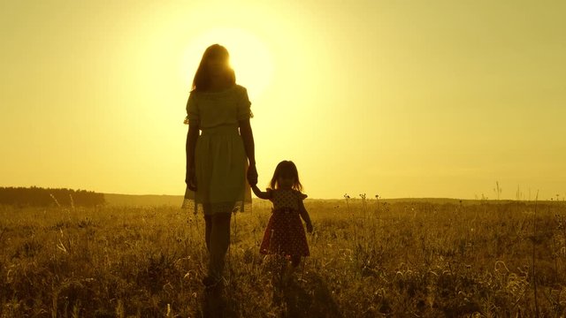 Mom walks on field with her little daughter in sunset. baby holds mom's hand, happy family walks in evening out of town. mother and baby are resting in park. child plays with mother in meadow.
