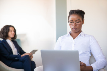 Fototapeta premium Focused young female professional working in office lounge. Diverse business women sitting in armchairs, using smartphone and laptop in office lobby. Workplace concept