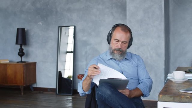 Medium shot of grey-haired bearded man sitting at desk in relaxed pose, analyzing document and making amendments while enjoying music using headphonesMedium shot of grey-haired bearded man sitting at 