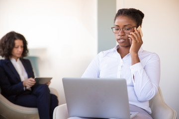 Confident young female manager calling on cellphone at computer. Black business woman sitting in armchair, using laptop and talking on phone in office lobby. Communication concept