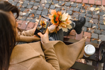 woman sitting in city park drinking coffee taking picture of bouquet of maple leaves