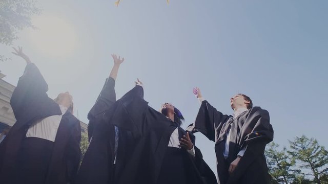 Group of multi ethnic graduates in traditional clothes tossing their academic caps up in the air happily on the blue sky background. University graduation, success. Slow motion.
