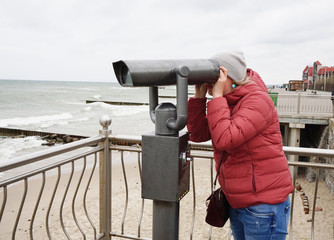 A young woman in a jacket and jeans looks through a public telescope on the waterfront and watches...