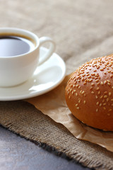 A sesame bun and a mug of coffee on kraft paper and burlap tablecloth.