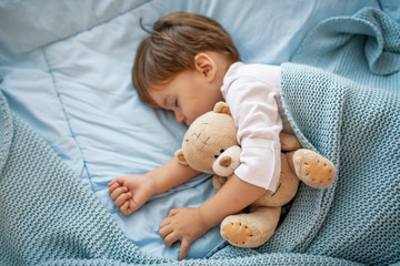 Adorable kid boy after sleeping in grey bed with toy.  Cute healthy little toddler baby boy child sleeping / taking a nap under blanket in bed while hugging teddy bear. - obrazy, fototapety, plakaty