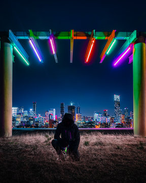 A man kneeling below a neon overhand with a city in the bacgkground.