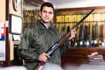 Confident hunter male wearing windbreaker in hunting shop with rifle in hands