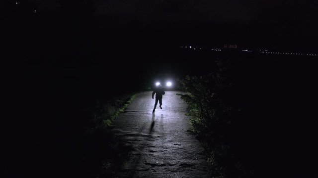 Young man dancing front car with lighting headlight on dark road in night. Drone view man jumping on dark road front illuminated car headlights