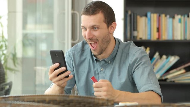 Excited man buying online with credit card and smart phone in a coffee shop