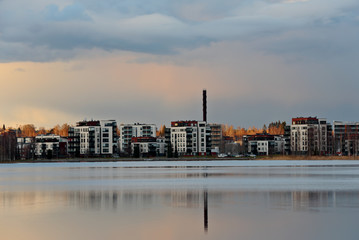 A city scape with new white apartment houses on the lake shore. Setting sun tints the cloudy sky