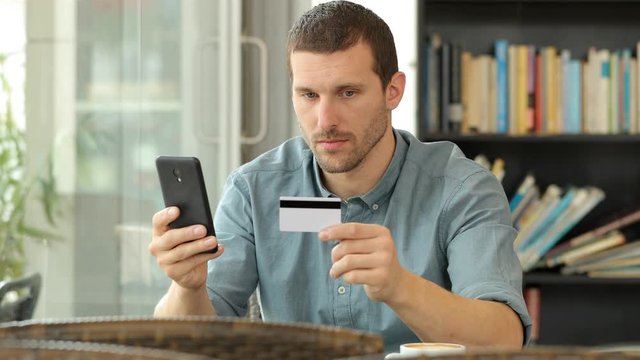Serious man using a credit card and smart phone to buy on line in a coffee shop