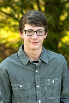 Teenage boy with glasses standing outside in front of a tree wearing a gray button up long sleeved shirt and denim jeans