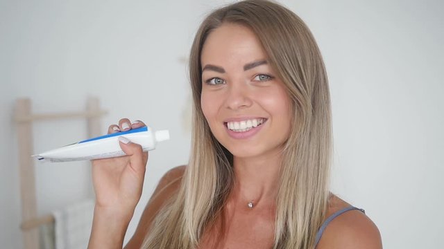 Beauty brushes her teeth in a modern bathroom. Attractive young woman raises a tube of toothpaste and smiles. Toothpaste advertisement. Beautiful smile, healthy teeth