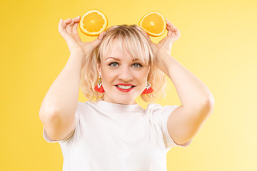 Cropped view of sunny blonde in white shirt looking at camera while posing with fruits on yellow isolated background. Happy girl keeping oranges and smiling. Concept of happiness and summer.