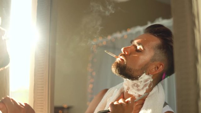 Handsome man looking in the mirror, smoking a cigarette and shaving his beard with a straight razor