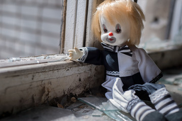 Mystical scary harlequin doll on the windowsill of an abandoned building. Points a hand towards the window. Abandoned children concept.