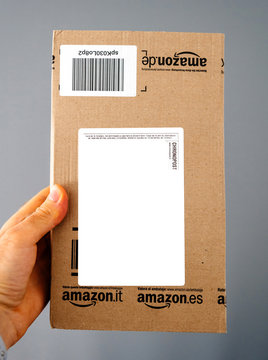 PARIS, FRANCE - APR 24, 2018: Man holding the front view with white sticker of Amazon prime cardboard delivery parcel box against gray background - on-line e-commerce shopping through Chronopost post