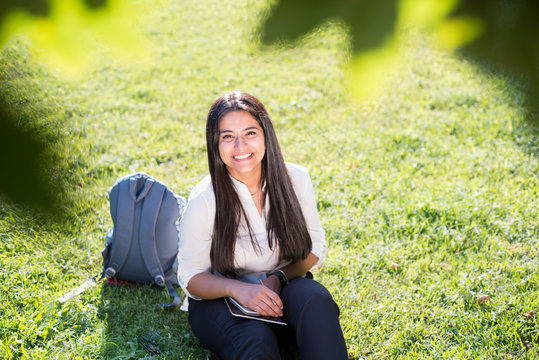 Indian girl, student, smiles, sits on the green grass, with a backpack and notepad, writes
