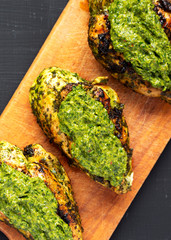 Grilled chimichurri chicken breast on a rustic wooden board on a black surface, top view. Flat lay, overhead, from above. Close-up.