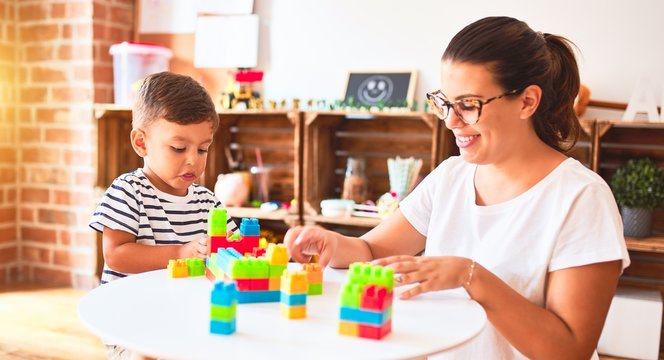 Beautiful teacher and toddler boy playing with construction blocks bulding tower at kindergarten
