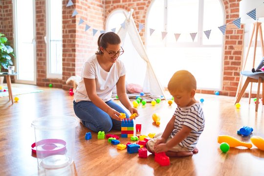 Beautiful teacher and toddler boy playing with construction blocks bulding tower at kindergarten