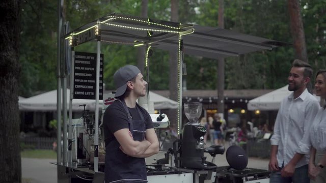 Barista man selling coffee to young couple in the movable coffee shop on the street