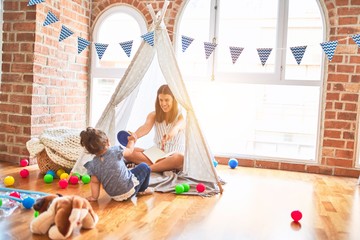 Young beautiful teacher reading book to toddler inside tipi at kindergarten
