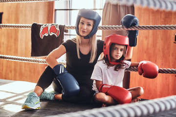 Young cute girl in helmet and her beautiful female boxing trainer are posing for photographer on the ring.