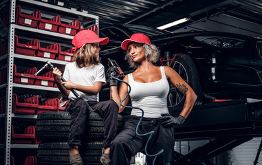 Fototapeta na wymiar Smiling woman and her little cheerful helper are posing for photographer at dark auto service as great team.