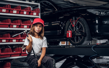 Shelf with tools, shiny car and little girl in hat with big wrench in hands.