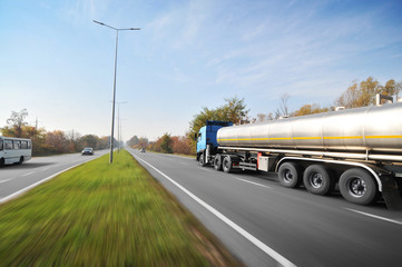 Big metal fuel tanker truck shipping fuel  with other cars on the countryside road in motion with trees against blue sky