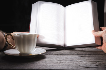 Young man is sitting on a sofa and reading a book while holding a paper cup