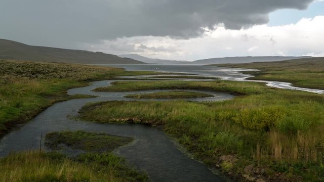 Time lapse of dramatic storm blowing over meadow with river winding into lake as the sun comes out after the darkness.