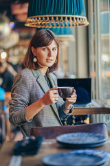 Beautiful girl in gray blazer, drinking coffee and working on net-book   in cafe. Freelancer working in coffee shop. Learning online.