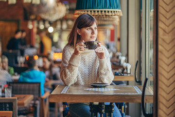 Beautiful young woman in warm sweater in the cafe near the window with coffee. Cozy autumn or winter morning.