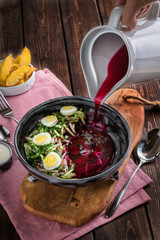 Hand pours from a white jug ?old beetroot soup with eggs. Baked potatoes, souce, pink tablecloth on the rustic wooden background. Top view