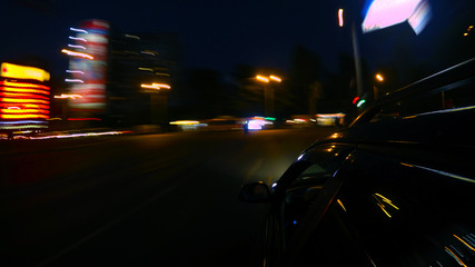 A dark-colored car is moving rapidly along the illuminated street of a night city, on a blurred abstract background a part of the car with a mirror is visible 