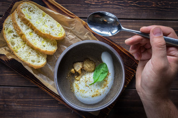 Man's hand holding spoon over a cappuccino soup with mushrooms on wooden cutting board. Three slices of white bread on the brown wooden background in restaurant. Horizontal photo.
