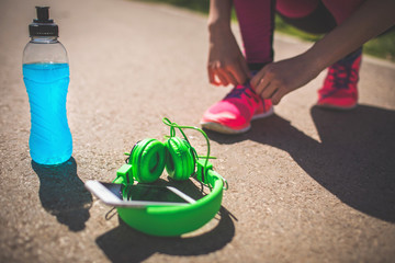 Young Fitness girl tying shoes before start running. Female runner with energy drink and headphones on the track. Sport recreation concept. Healthy lifestyle.